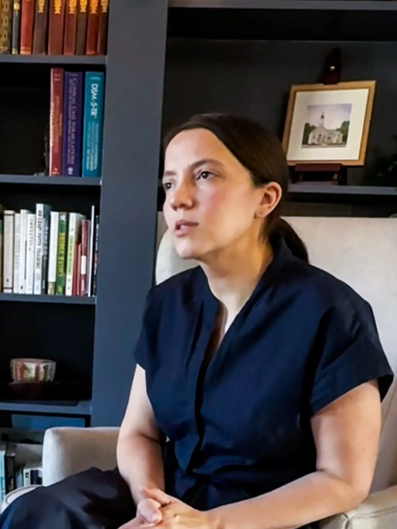 A woman with dark hair tied back, wearing a black shirt, sitting on a beige chair in a room with a bookshelf and framed picture on a dark shelf behind her.