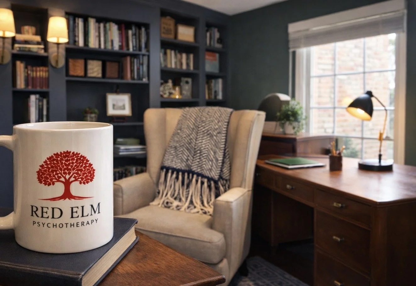 A cozy psychology office with a wooden desk, a beige armchair with a gray and white throw blanket, a bookshelf filled with books, a window with blinds, and a coffee mug that says 'Red Elm Psychotherapy' on a book on the desk.