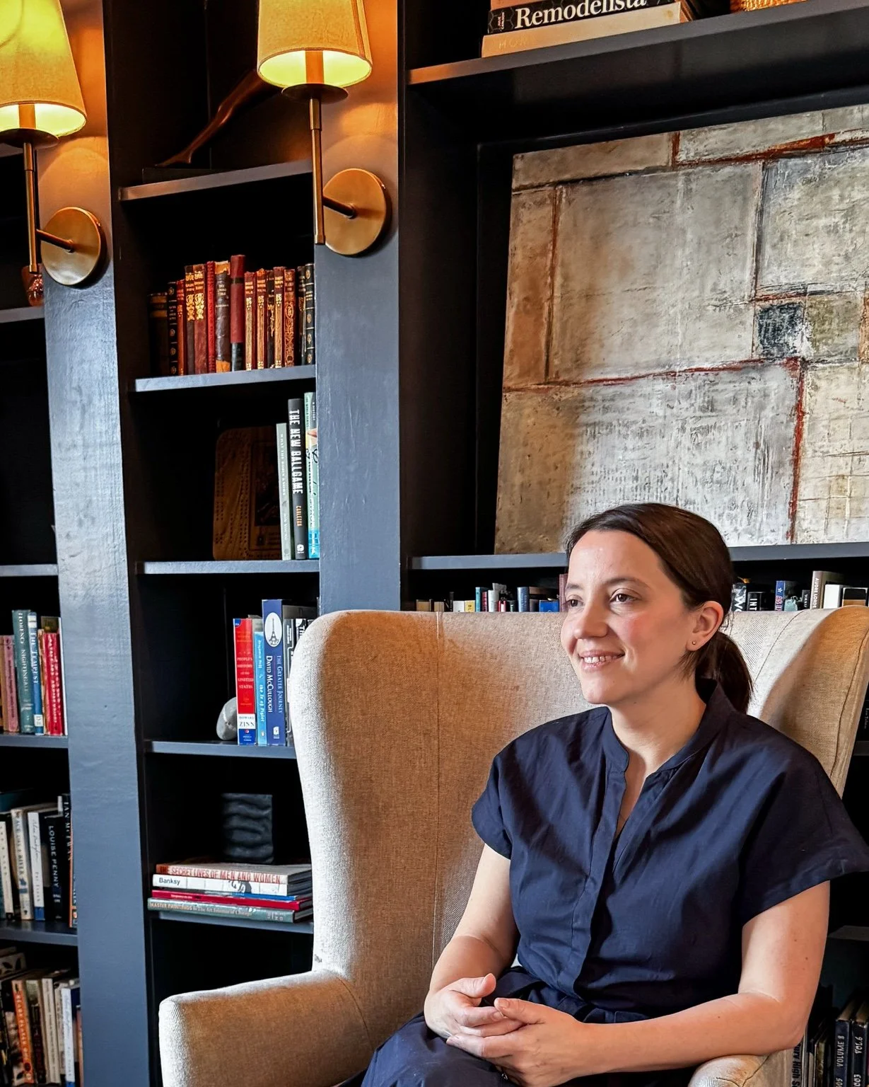 A woman sitting in a beige armchair in a bookshelf-lined room, with warm lighting from lamps and a painting or textured wall behind her.