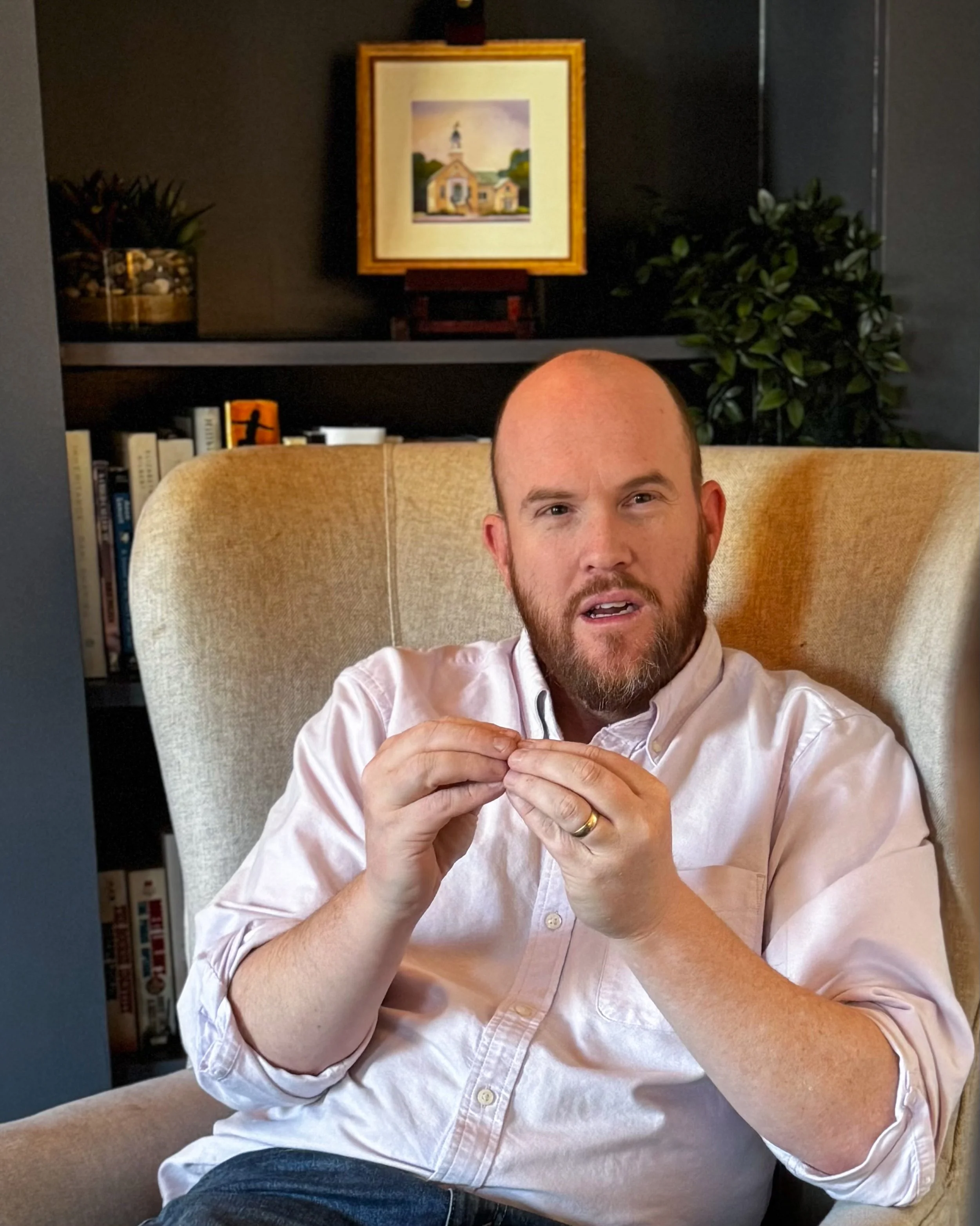 A man with a beard and a bald head sitting in a beige armchair, wearing a white button-up shirt, with a bookshelf and framed picture in the background.