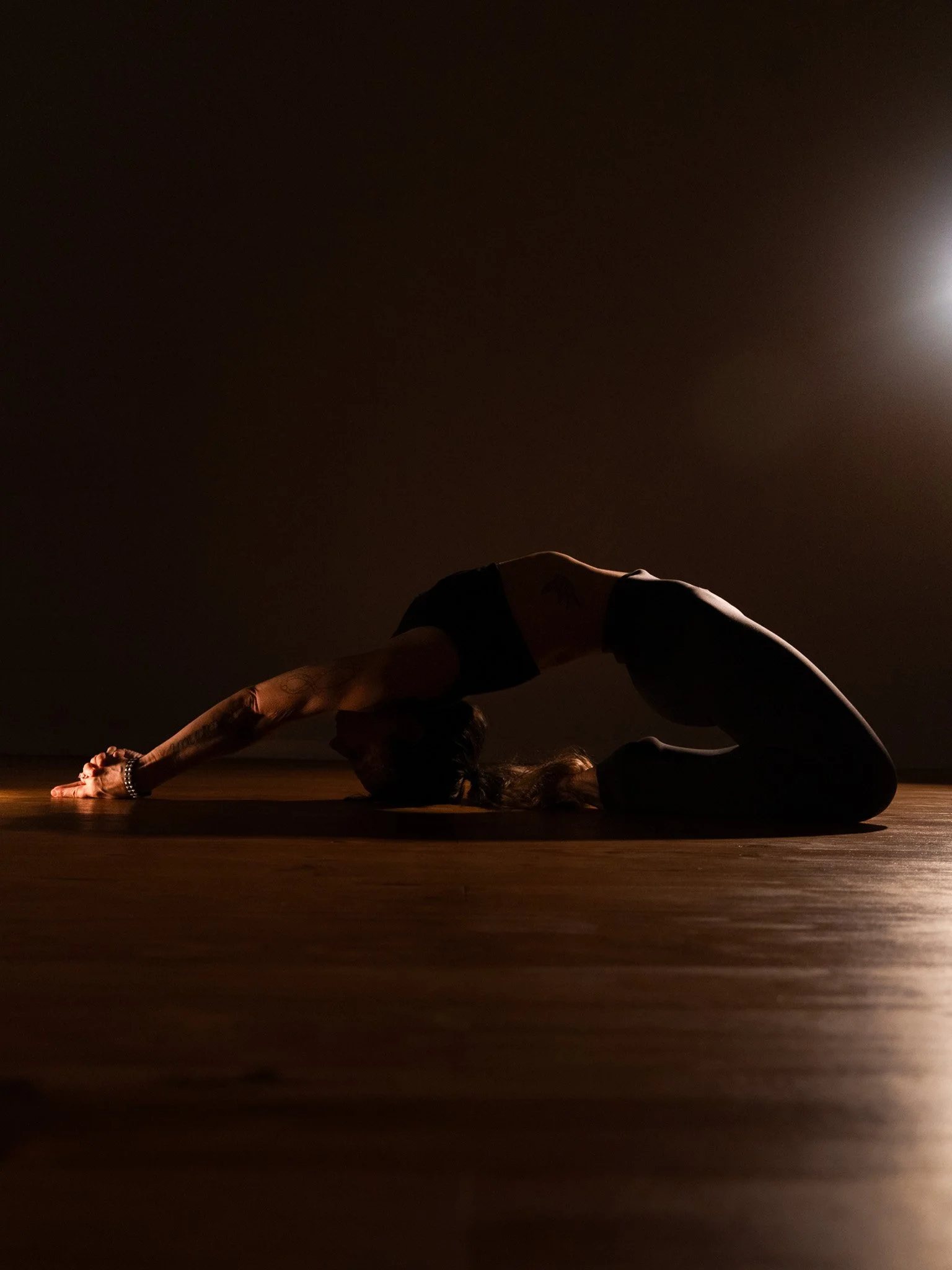 A person practicing yoga in a dimly lit room, performing a forward bend pose on a wooden floor, with a bright light source in the background.