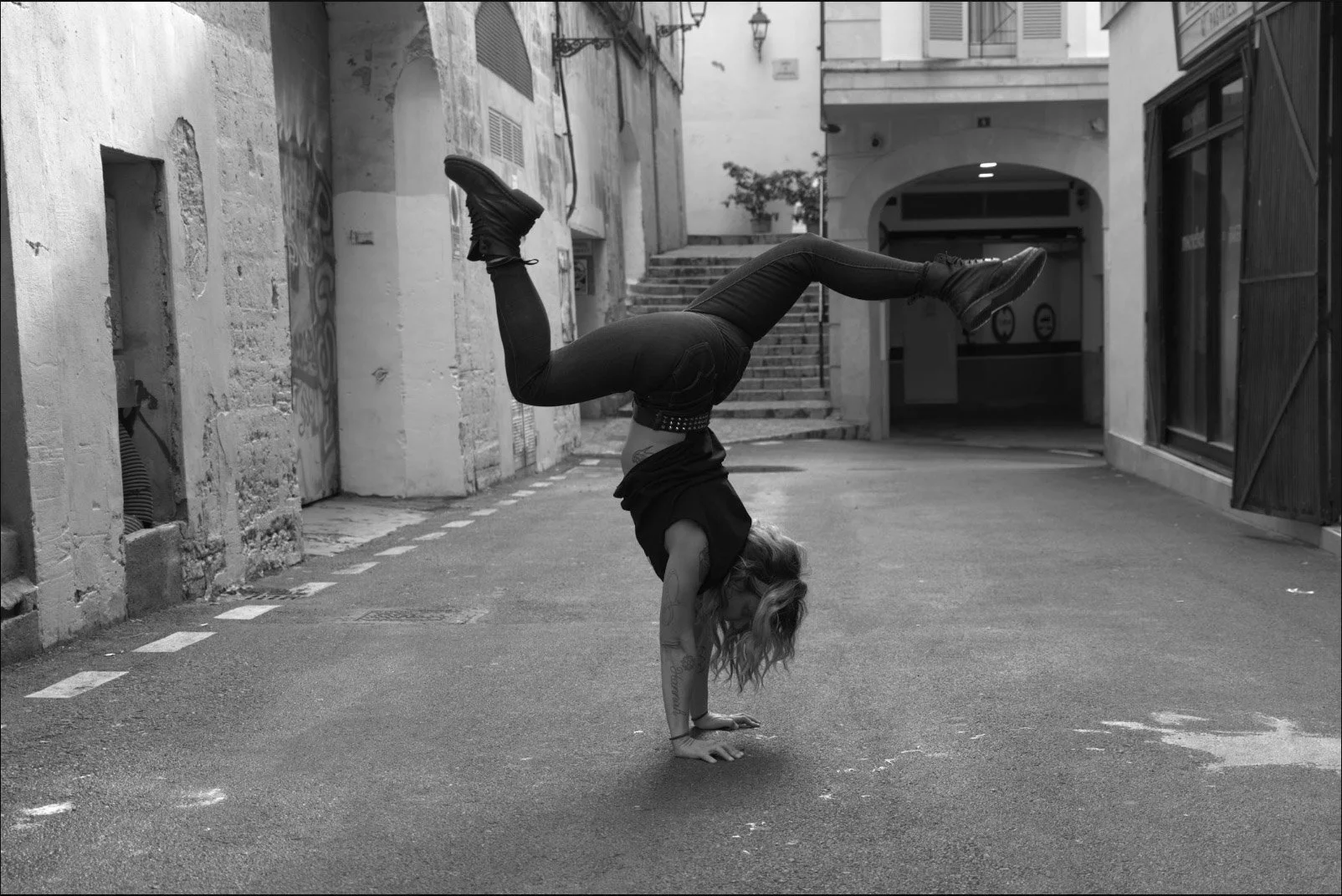 A person with long hair and tattoos performing a handstand with legs bent backward on an urban street in black and white.