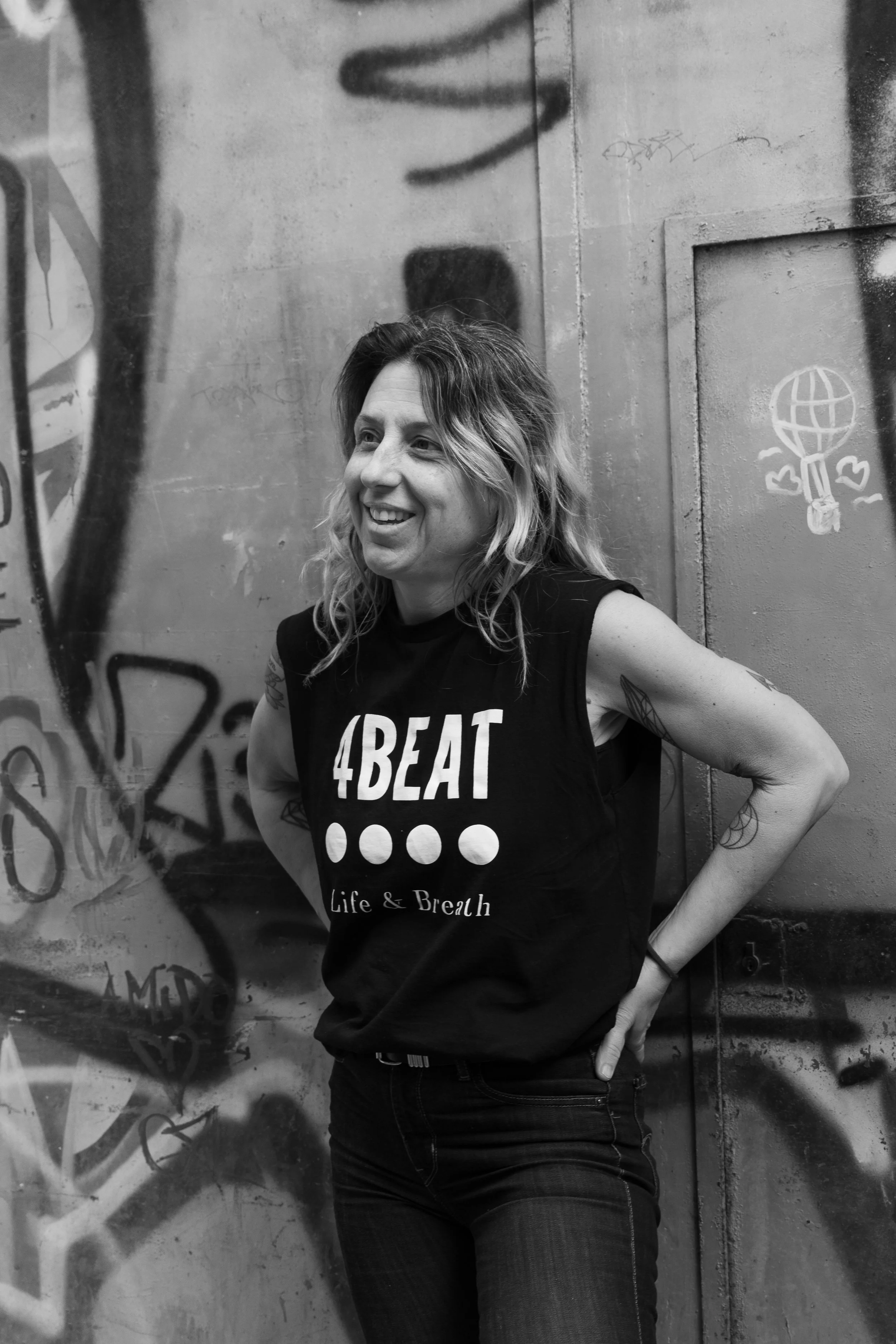 A woman with wavy hair standing against a graffiti-covered wall. She is smiling, wearing a black sleeveless shirt with the text '4BEAT' and additional smaller text 'Life & Breath', and dark jeans.
