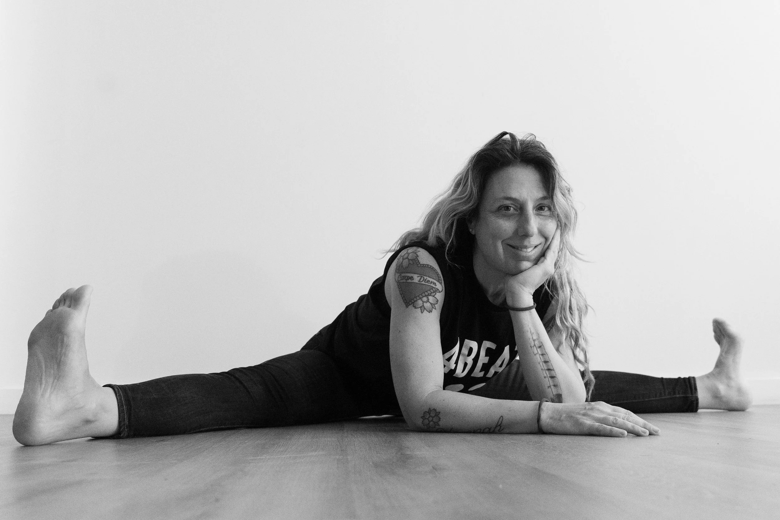 Black and white photo of a woman doing a yoga pose on the floor, lying face down with one arm bent under her head, and the other arm extended in front of her. She is smiling and has tattoos on her left arm and thigh. Her hair is wavy and shoulder length.