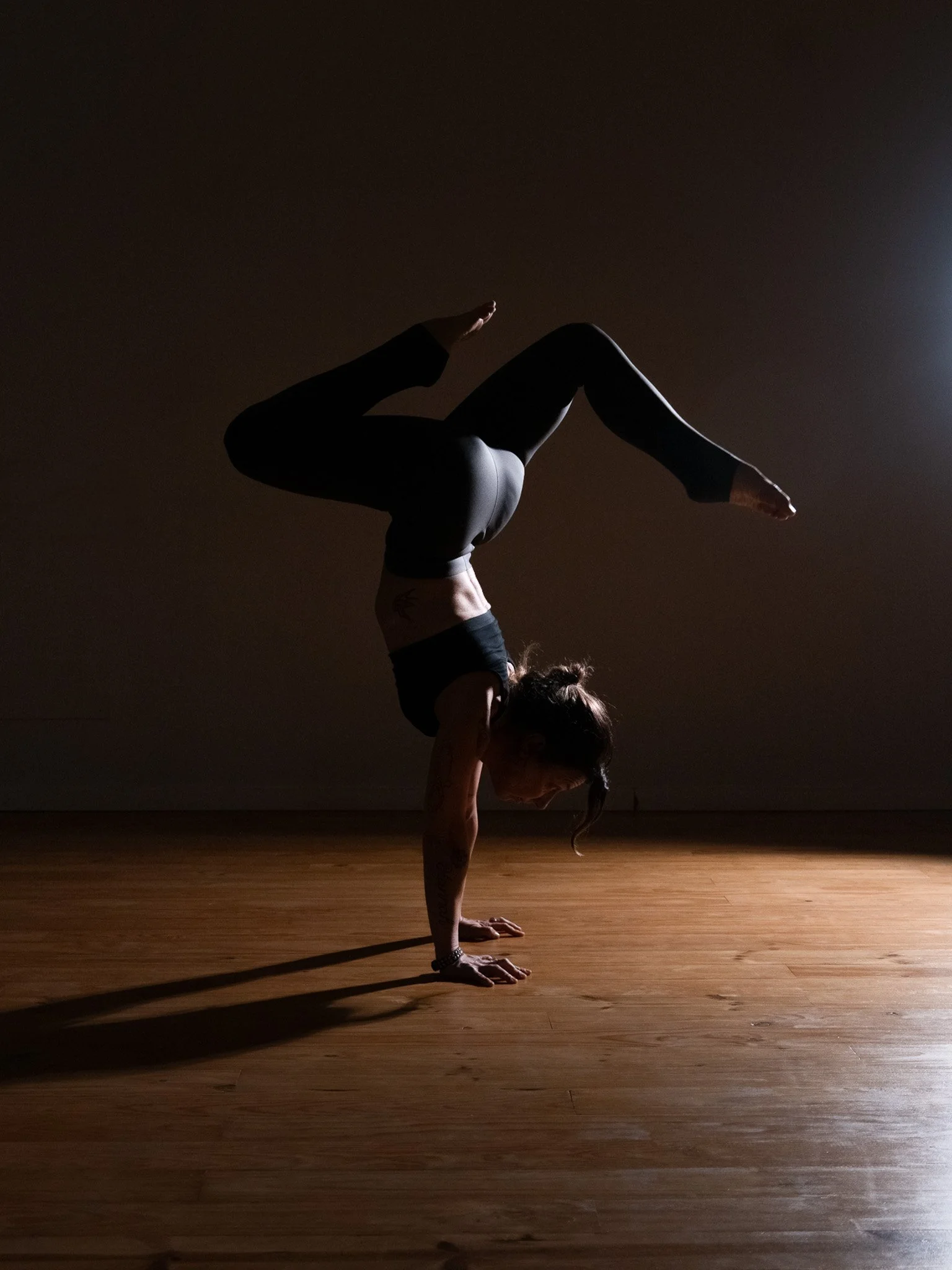 A woman in yoga leggings balances upside down in a handstand pose with her legs bent over her head in a dimly lit room with hardwood floor.