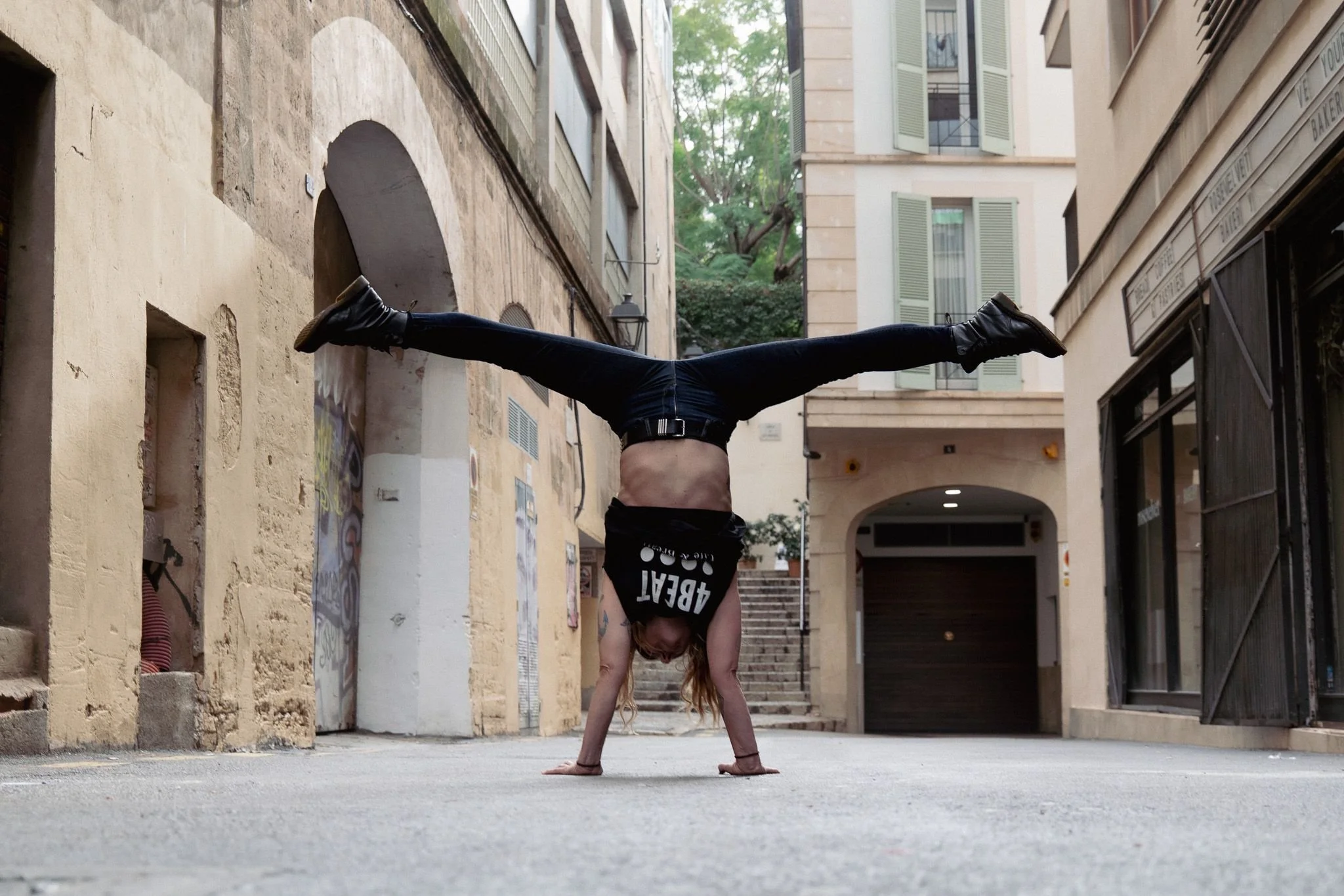 A woman with long hair performing a handstand in an alleyway, balancing on her hands with her legs spread wide apart, wearing a black shirt, black pants, and black shoes.