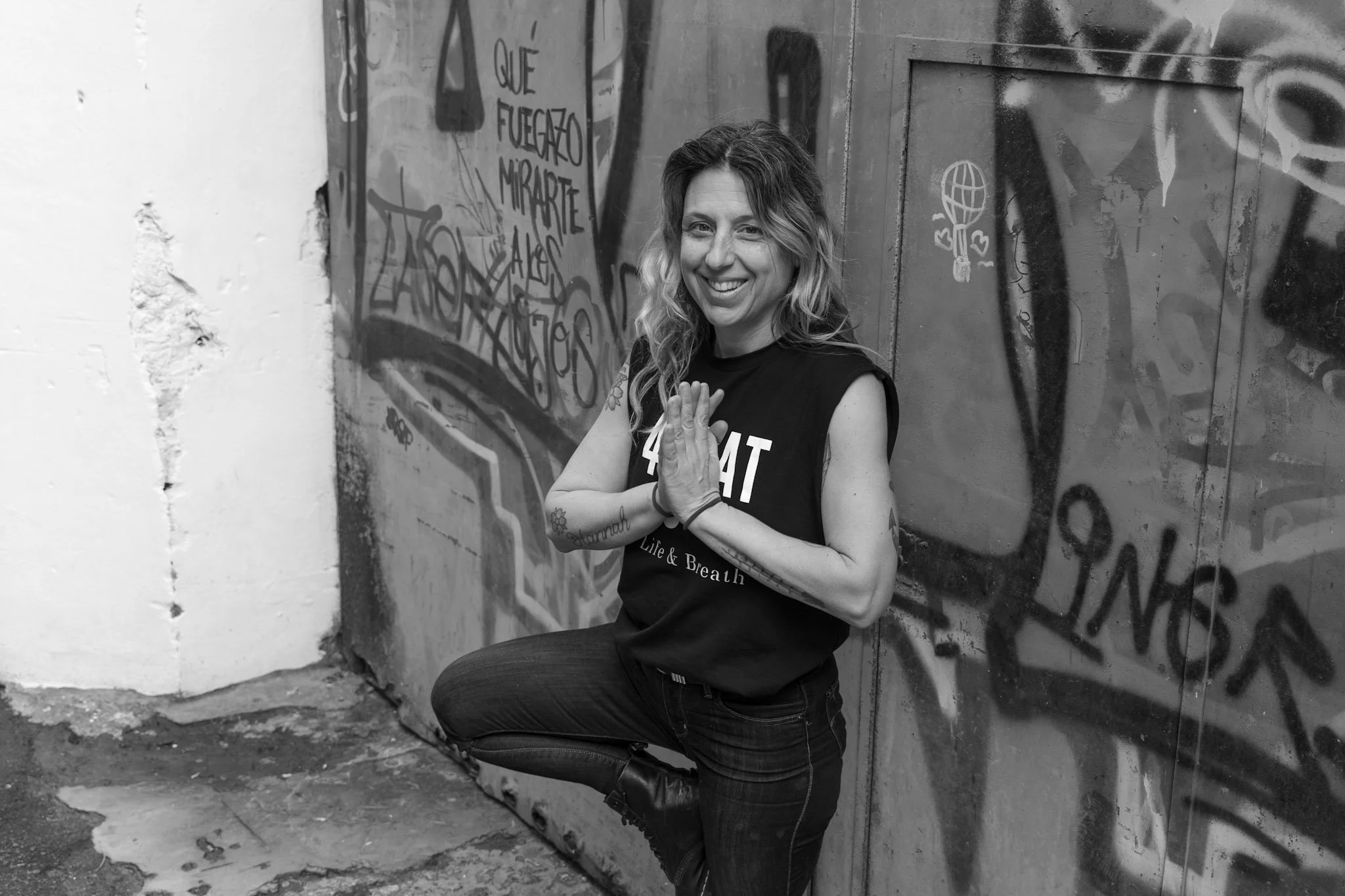 A woman with tattoos and wavy hair smiling, kneeling with hands pressed together in a prayer pose in front of a graffiti-covered wall.
