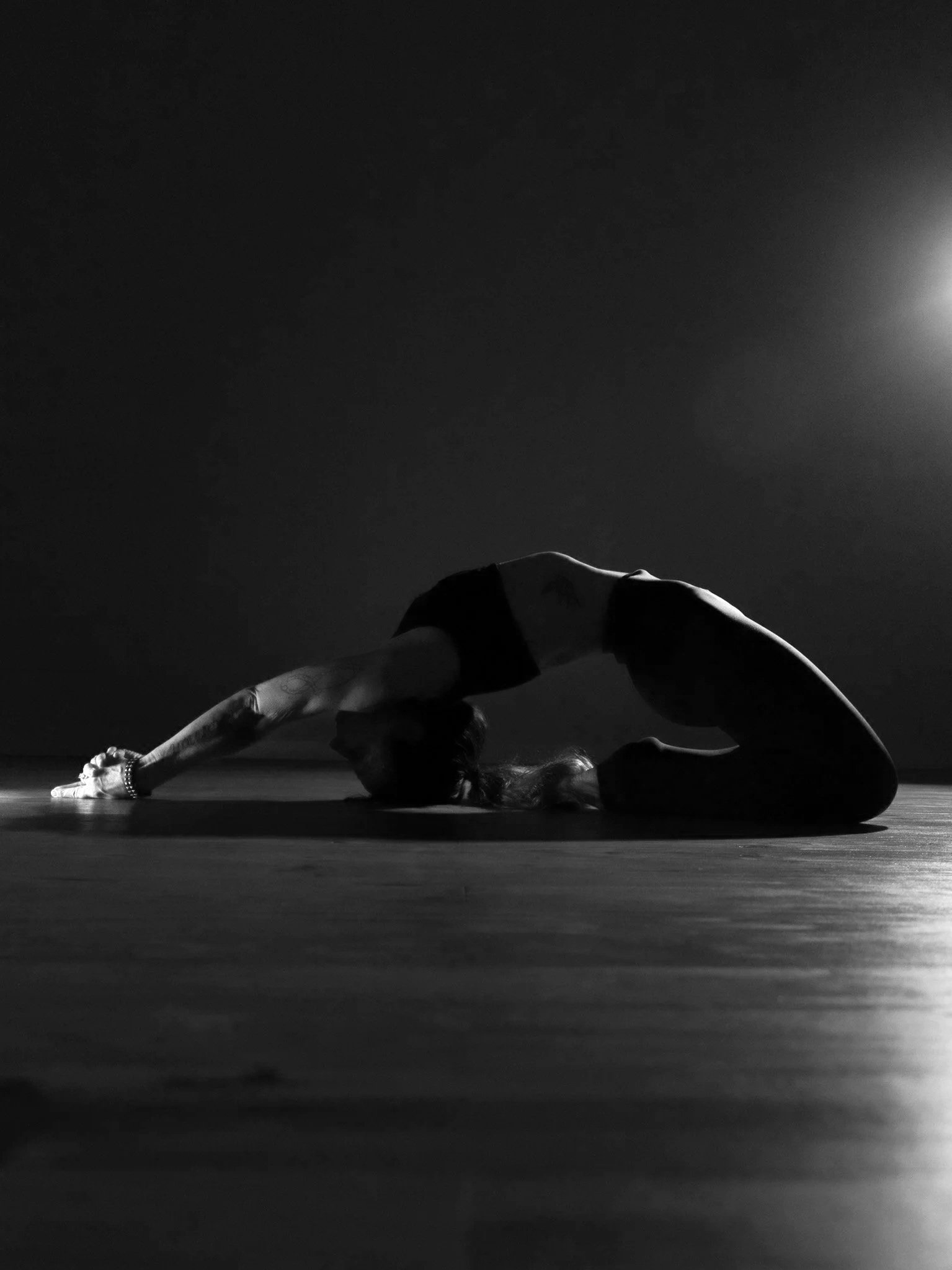 A woman performs a yoga pose on a dark floor in front of a bright light, with her body looking like a horizontal line, head resting on the floor, and arms stretched forward.