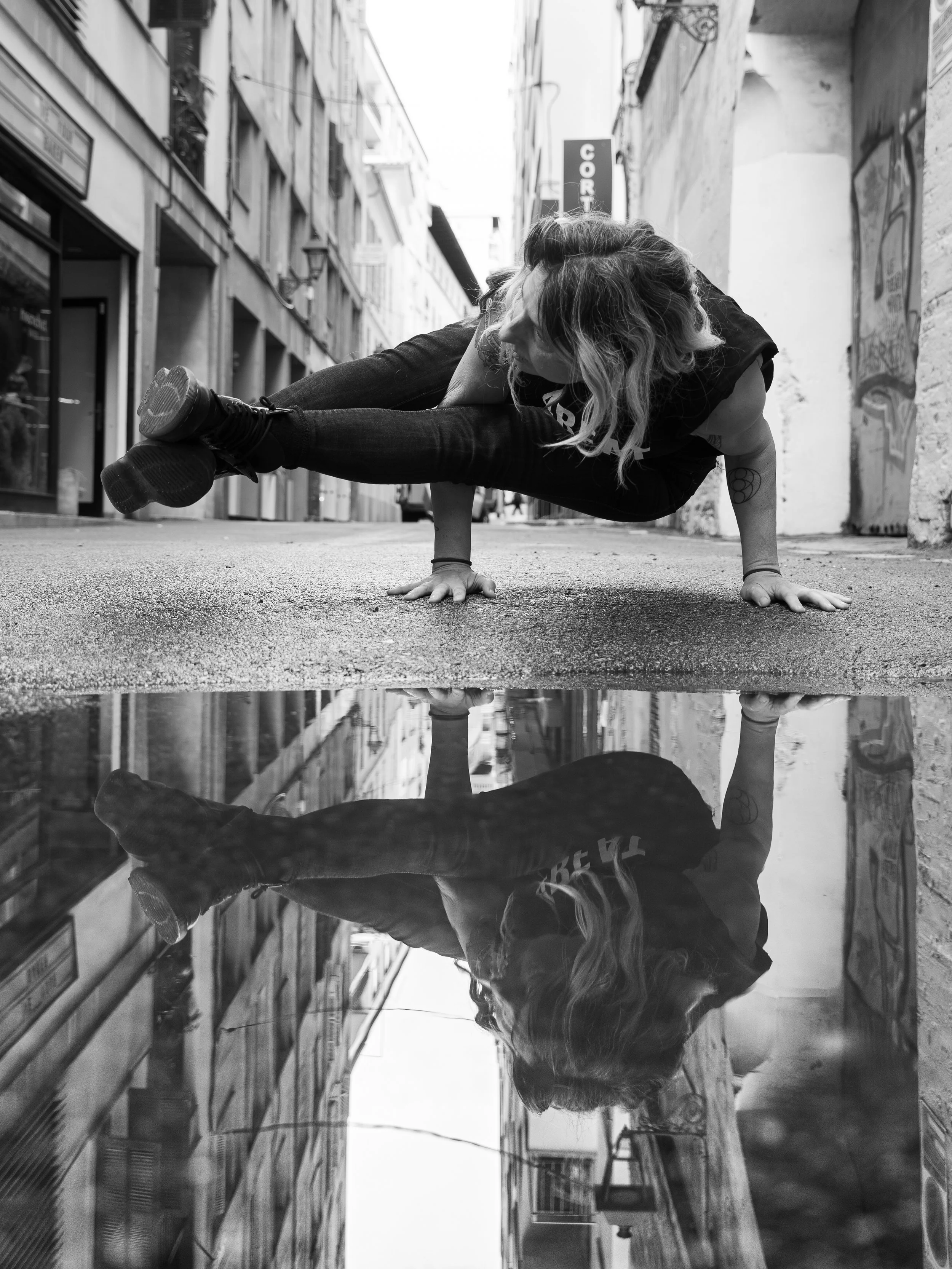 A person with curly hair performing a handstand on a city street, with reflection below showing the handstand, surrounded by buildings and a sky.