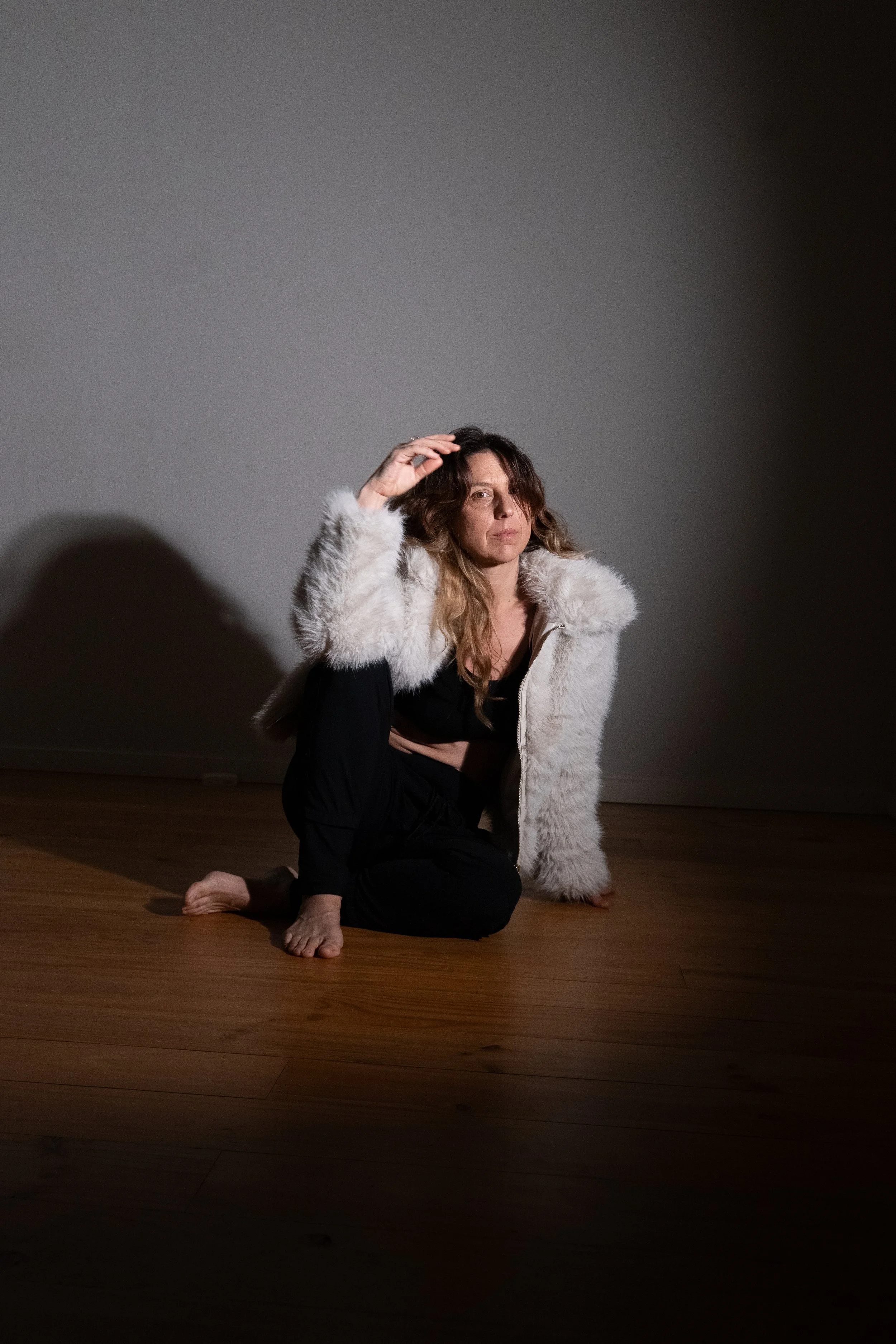A woman doing a yoga pose on a wooden floor against a plain gray wall, wearing a black outfit and a white fur jacket.