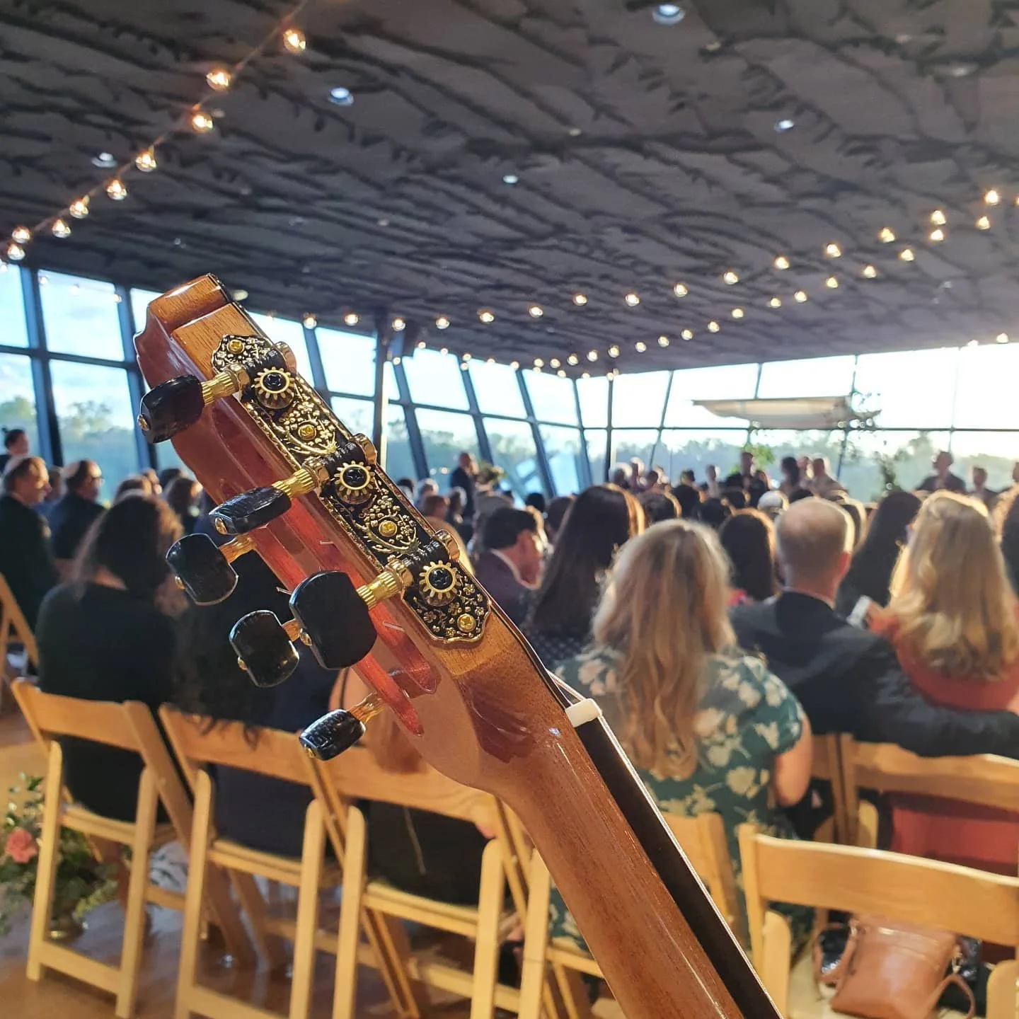 Close-up of a guitar headstock with tuning pegs, in an indoor concert venue filled with seated audience members during what appears to be an event or performance.