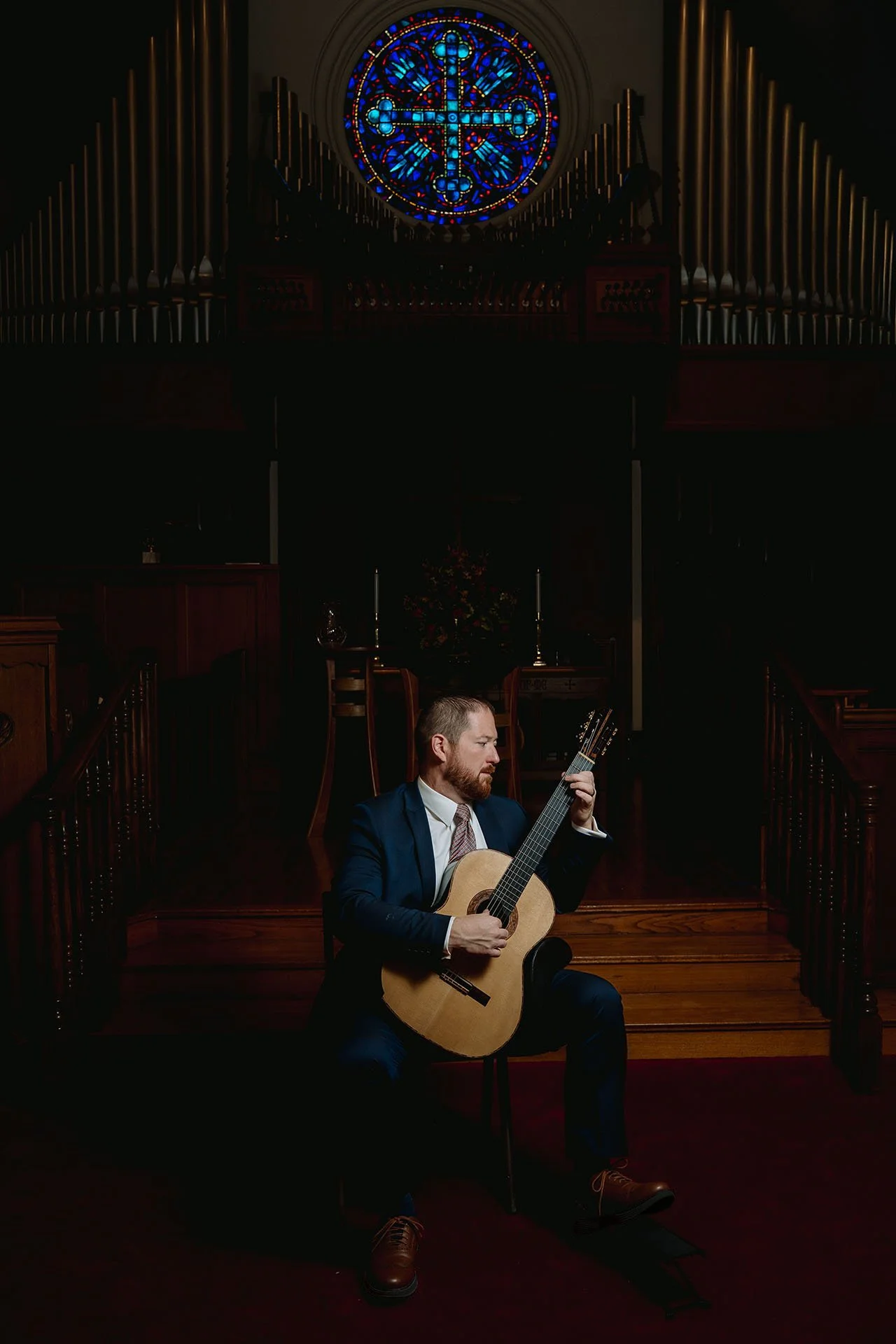 Dan Kyzer, Classical Guitar | Pictured is Dan Kyzer in a suit playing an acoustic guitar on a wooden church stage with a stained glass window and pipe organ in the background.