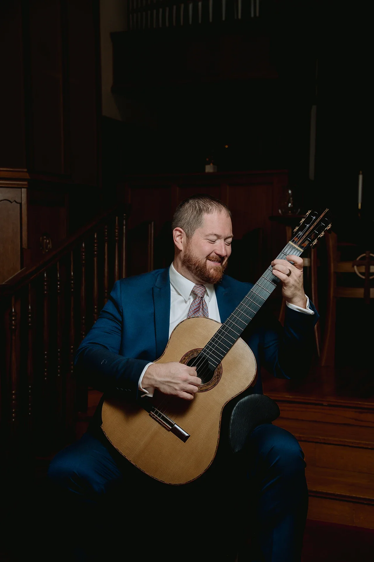 Dan Kyzer, Classical Guitar | Pictured is Dan Kyzer in a blue suit playing an acoustic guitar and smiling, sitting on a chair in a dimly lit room with wood-paneled walls and a staircase in the background.