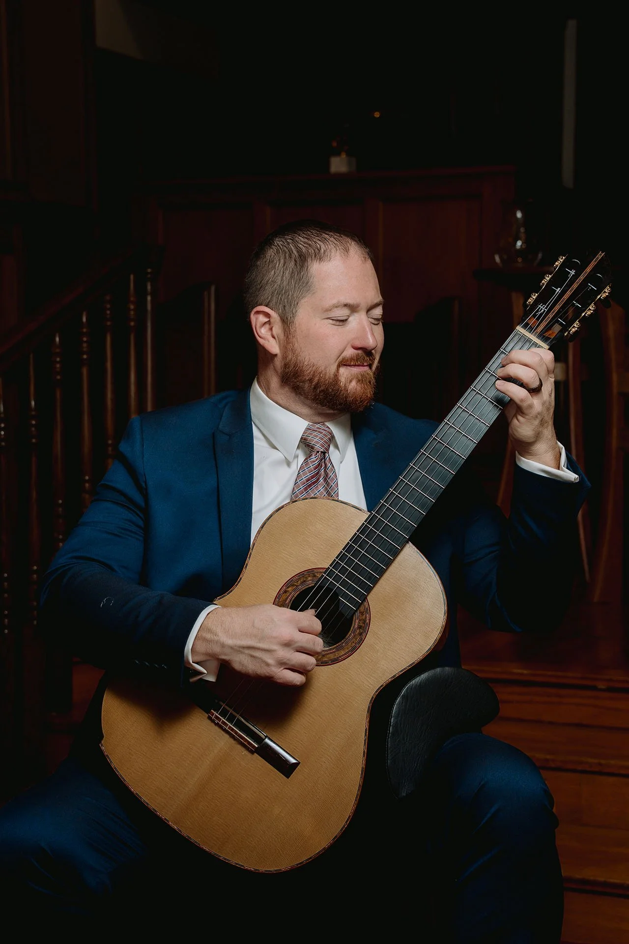 Dan Kyzer, Classical Guitar | Pictured is Dan Kyzer in a navy suit and tie is playing an acoustic guitar, sitting in a dimly lit room with wooden furniture.