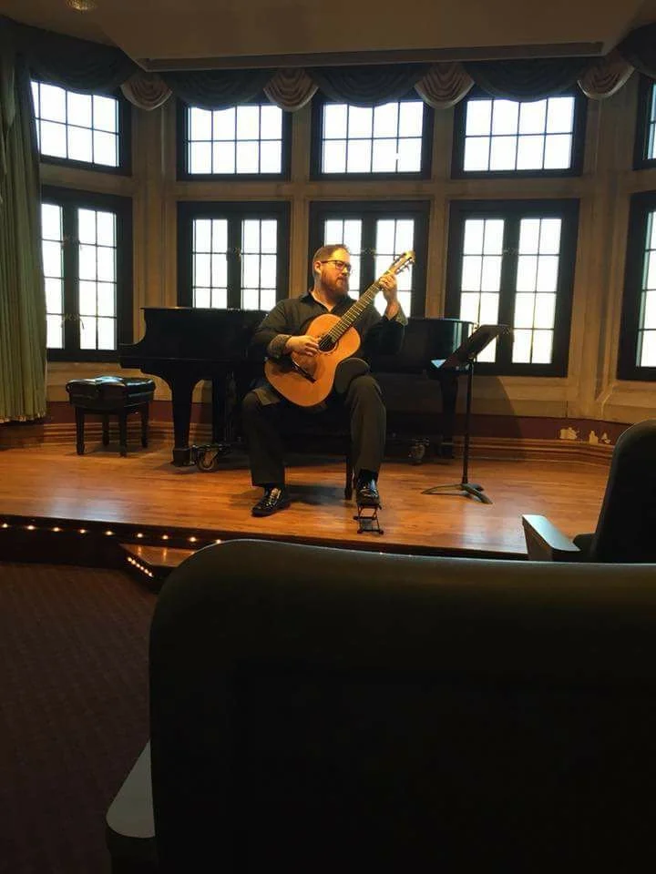 Dan Kyzer, Classical Guitar | Pictured is Dan Kyzer playing an acoustic guitar on a stage with a grand piano behind him, in a room with large window panes and decorative curtains.