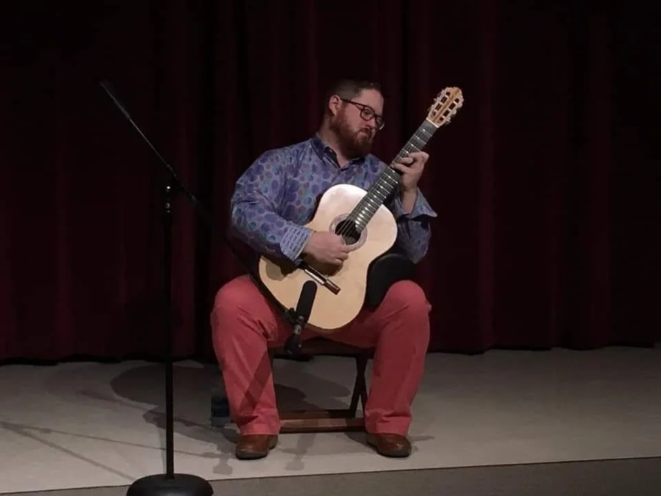 A man with glasses and a beard sitting on a chair playing an acoustic guitar on a stage with a dark curtain in the background.