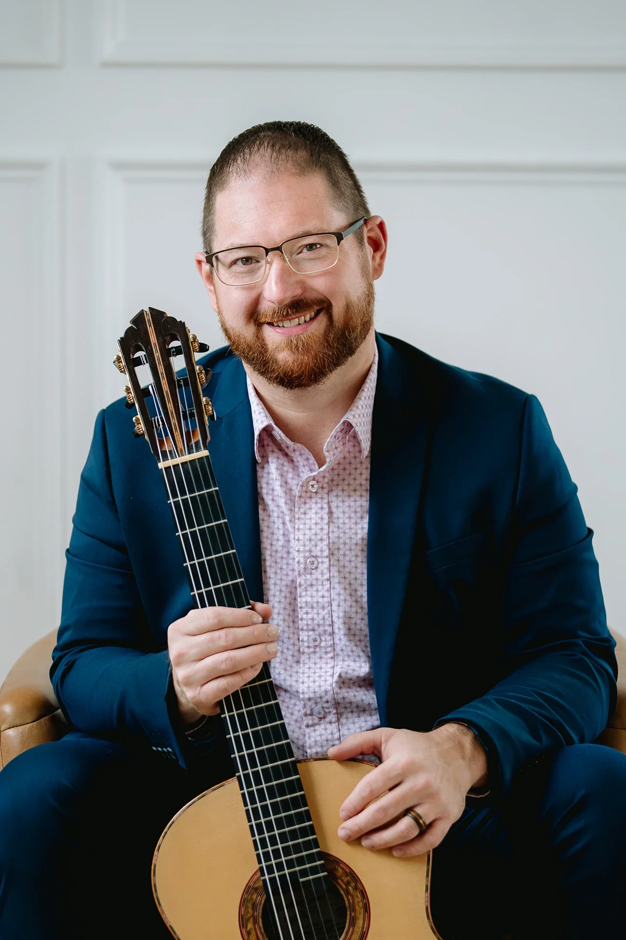Dan Kyzer, Classical Guitar | Pictured is a Dan Kyzer, wearing glasses in a blue suit holding an acoustic guitar indoors.