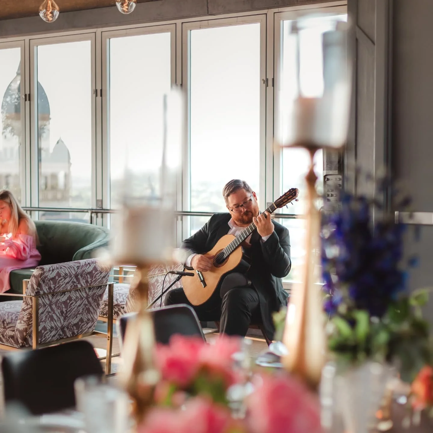 A man in a suit playing an acoustic guitar in a bright indoor space with large windows and a blurred foreground of flowers and decorative objects.