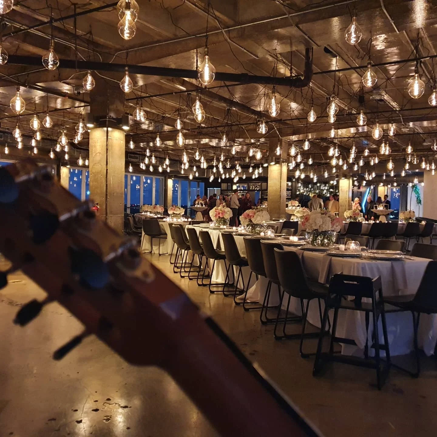 Dan Kyzer, Classical Guitar | Pictured is a banquet hall decorated with string lights hanging from the ceiling, tables covered with white tablecloths and floral centerpieces, surrounded by black chairs, with guests socializing in the background.