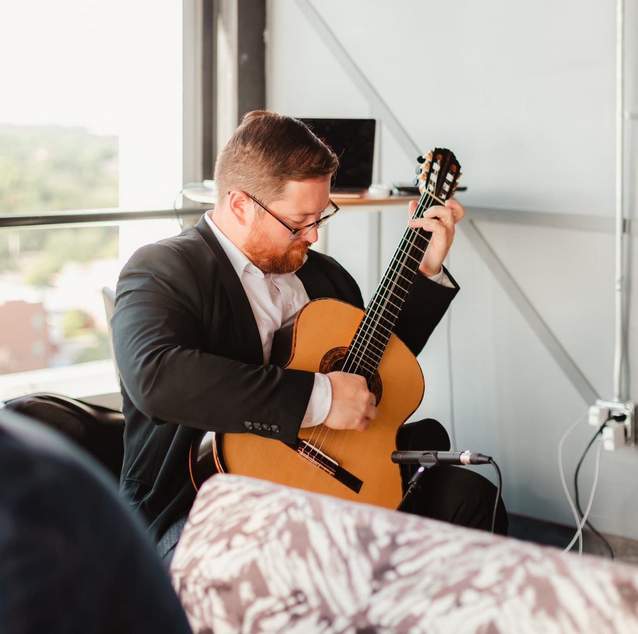 Man in business suit playing acoustic guitar indoors near a window.