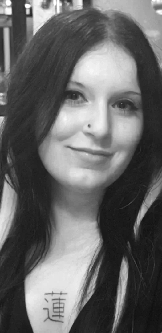 Black and white photo of a woman with long dark hair, smiling gently, with a tattoo of Japanese characters on her chest.