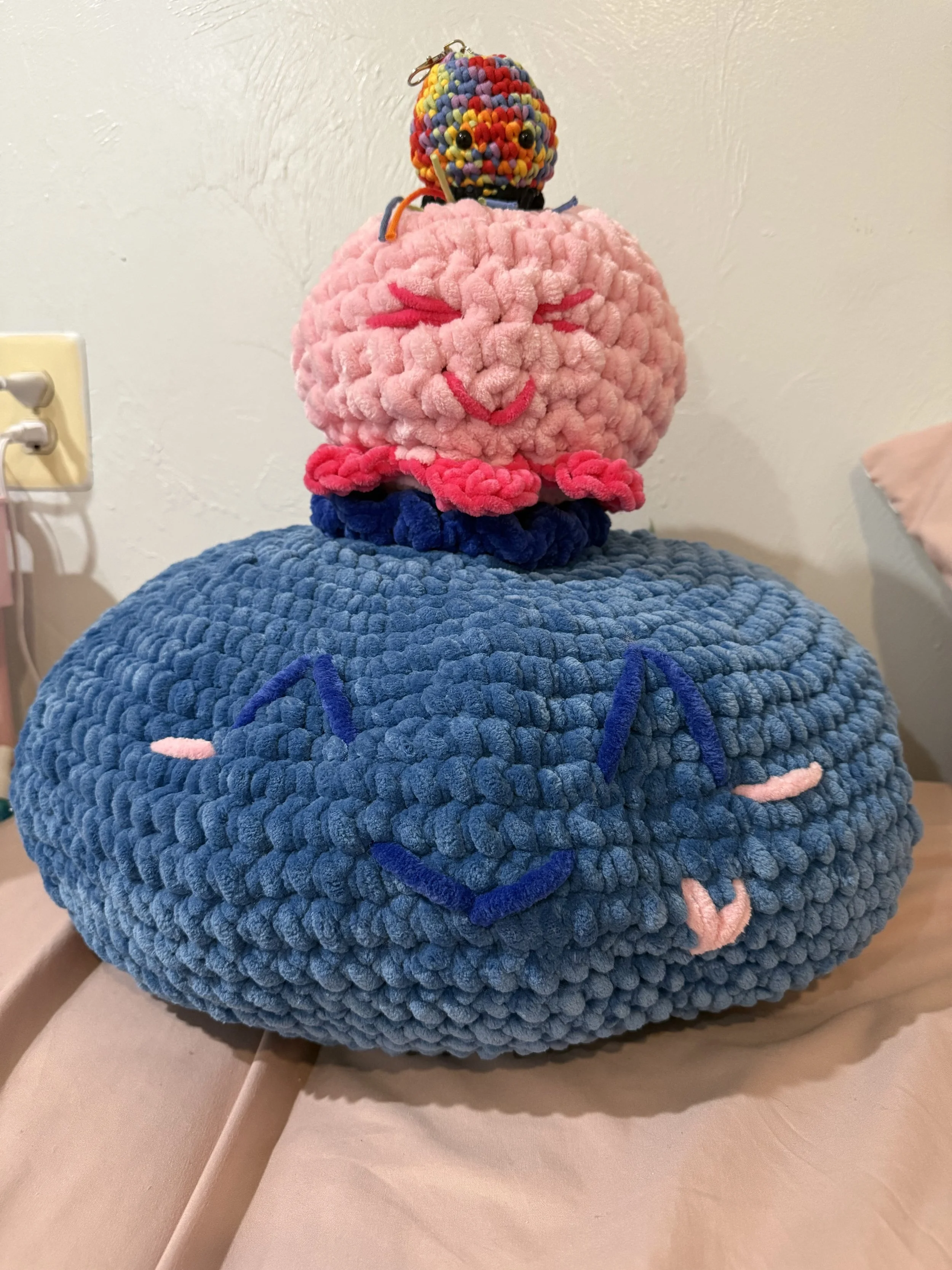 A plush pink and blue character with embroidered face and pink hands sitting on a beige couch.