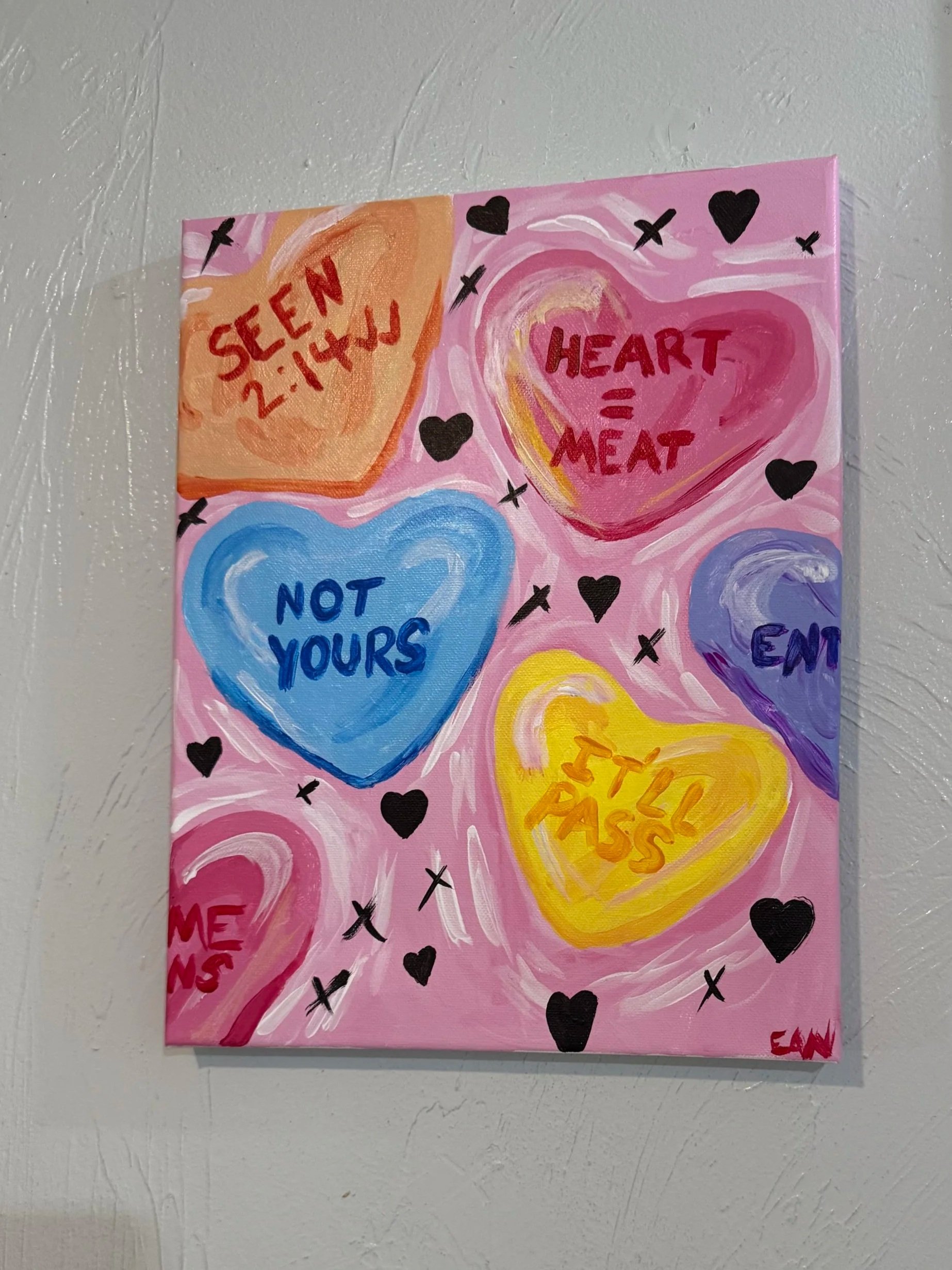 A colorful painting with hearts and words related to love and feelings. The paintings include phrases like 'HEART = MEAT,' 'SEEIN 2:14,' 'NOT YOURS,' and 'I FEEL.' The background is pink with various colored hearts and crosses.