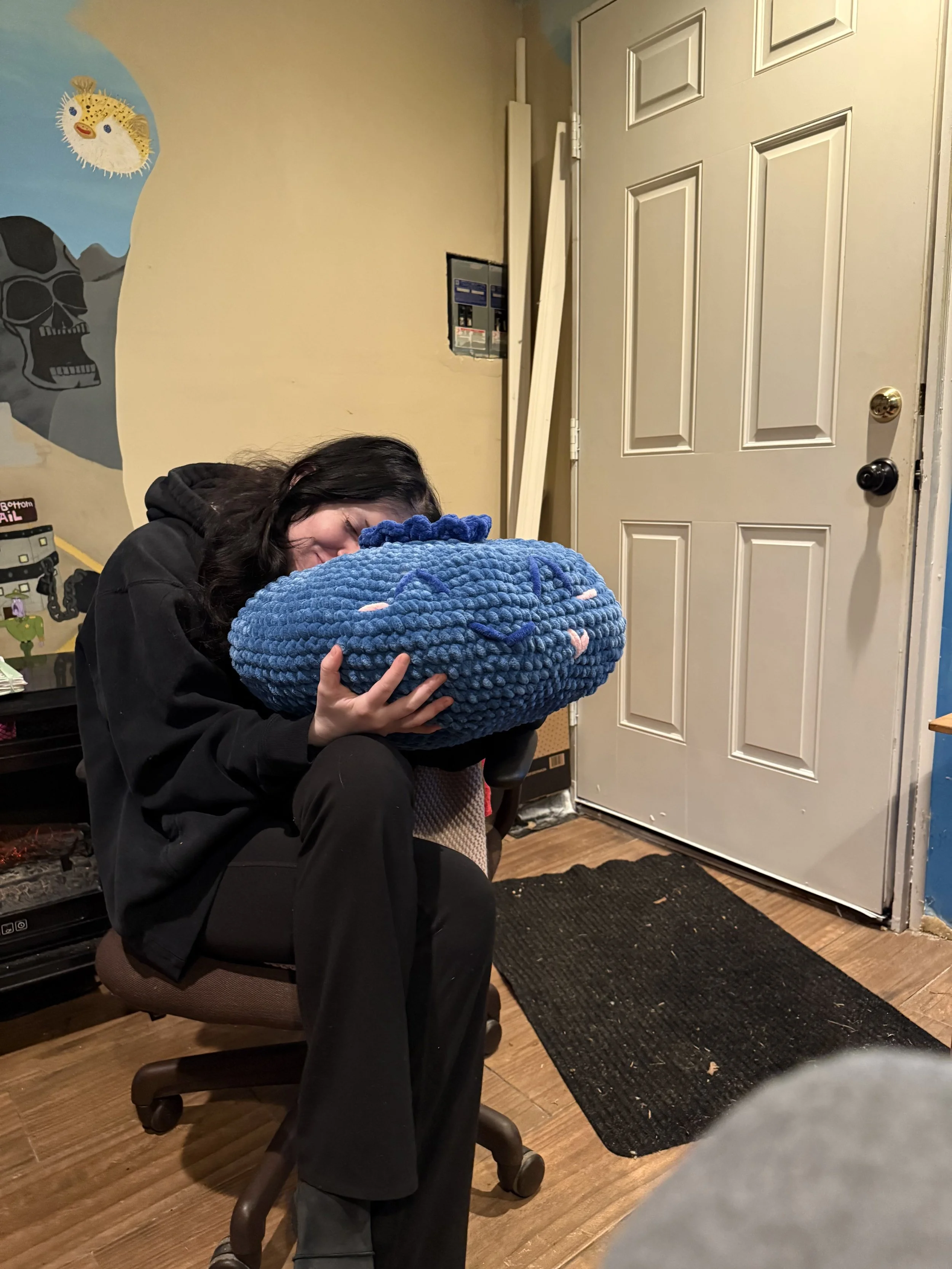 Woman with black hair smiling while hugging a large, plush, blue, textured stuffed animal in a room with yellow walls, closet door, and decorated wall with skull and fish artwork.