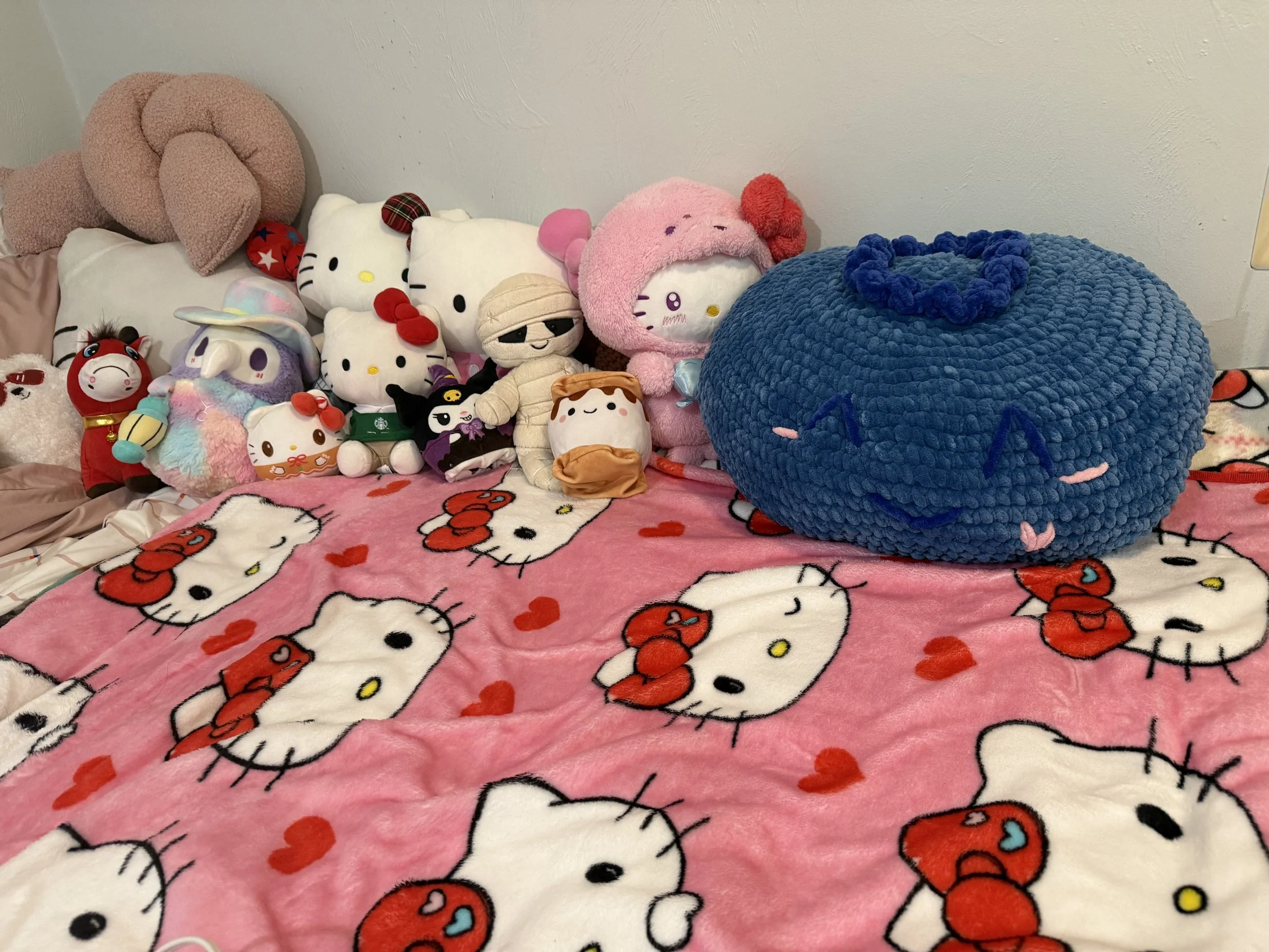 A collection of plush toys on a bed, with Hello Kitty plush toys and other stuffed animals, all on a pink Hello Kitty-themed blanket.