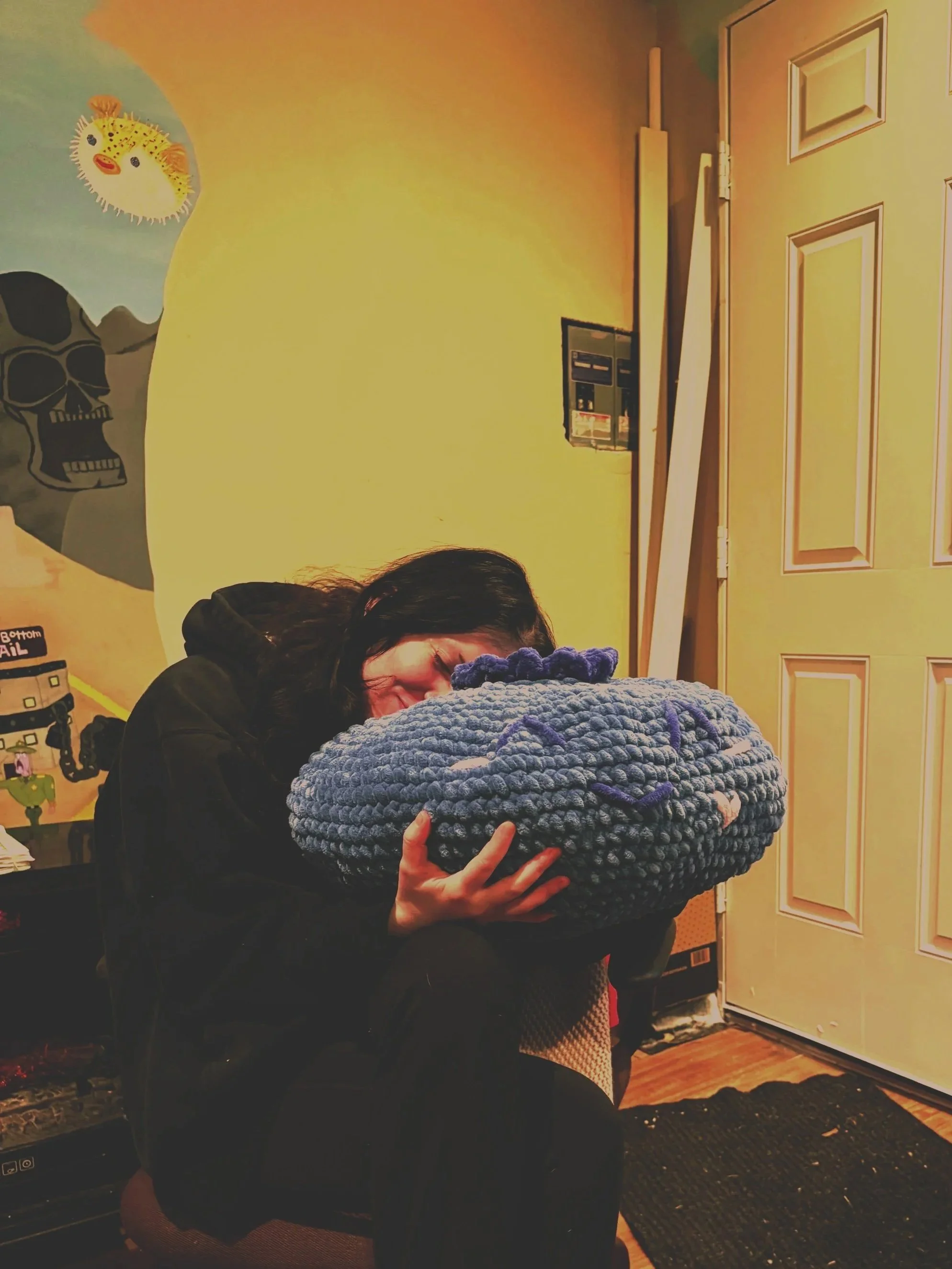 A woman with dark hair, wearing a black jacket, is hugging a large, chunky-knit, purple and gray blanket or pillow in a room with painted yellow walls featuring cartoonish skull and animal faces. A closed door and an open wall mailbox are visible in the background.