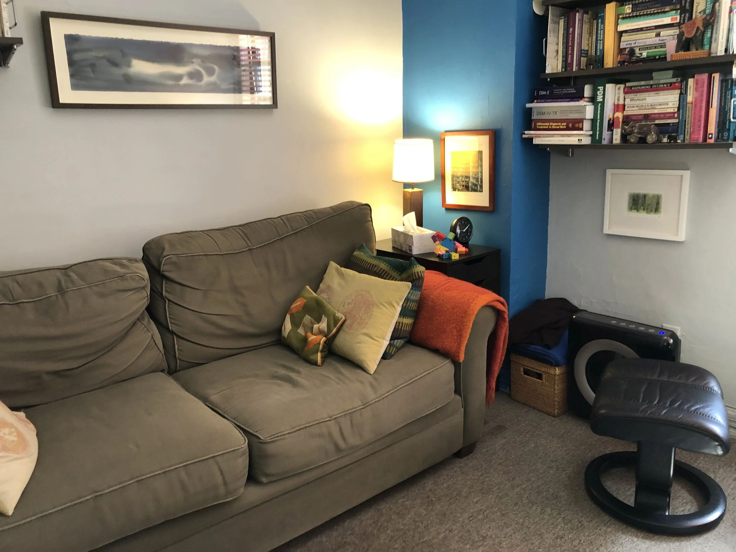 Living room with beige couch, colorful pillows, a small black table with a lamp, tissue box, and toys, a blue and white wall, bookshelf filled with books, a small framed picture, and a black ottoman on a beige carpet.