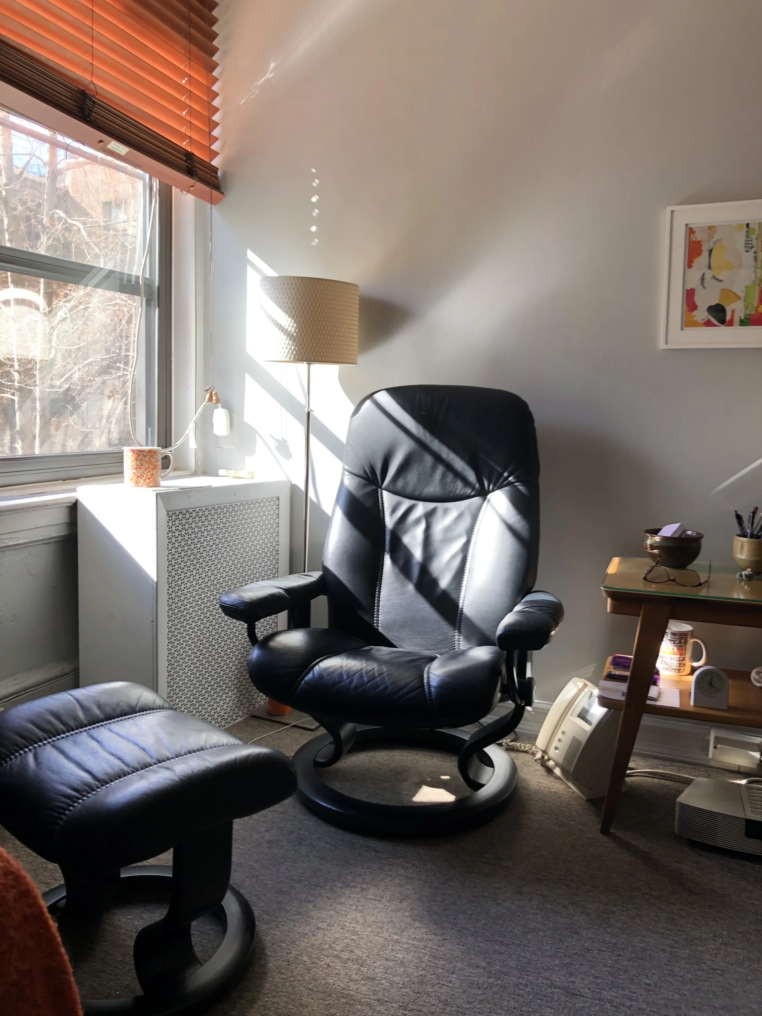 A sunlit home office corner featuring a black leather office chair and matching footrest, a floor lamp, a window with wooden blinds, a white radiator cover, and a small wooden side table with various items.