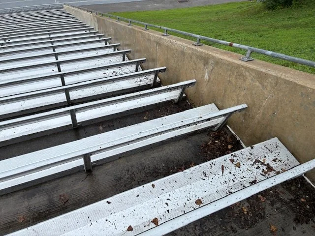 Empty metal bleacher seats with dirt and leaves, next to a concrete wall and a grassy area.