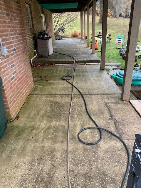A covered porch area with a concrete floor, a black garden hose and a gray extension cord lying on the ground, a brick wall on the left, and a backyard with a lawn, lawn chairs, and outdoor toys in the background.