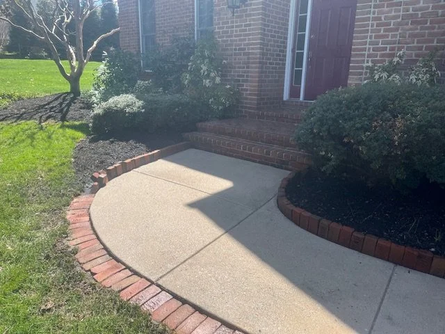 Paved concrete walkway with brick border leading to front steps of a brick house, surrounded by bushes and trees.