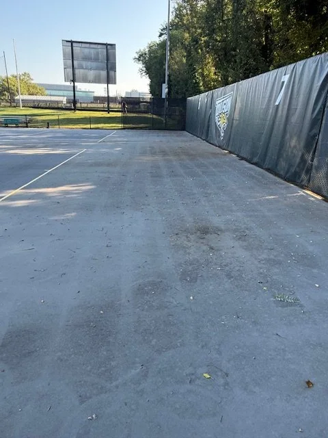 Empty outdoor tennis court with a black chain-link fence, a grey windscreen, and tennis court markings, with trees and a blue sky in the background.