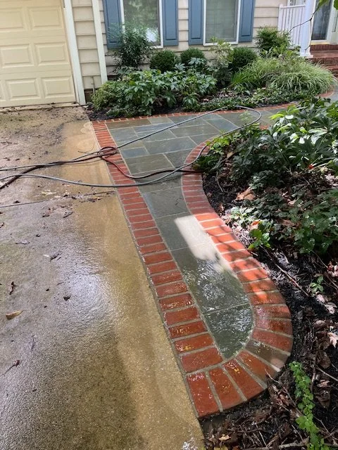 A partially completed sidewalk with stone tiles bordered by red bricks, next to a garden bed with green shrubs, near a house with beige siding and a garage.