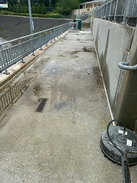 Sidewalk or pathway adjacent to a concrete wall, with wet puddles, a drain, and a floor cleaning machine.