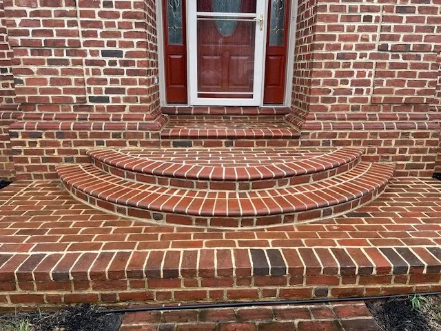 Brick front porch with three semi-circular steps leading up to a red front door with glass panels and a storm door.