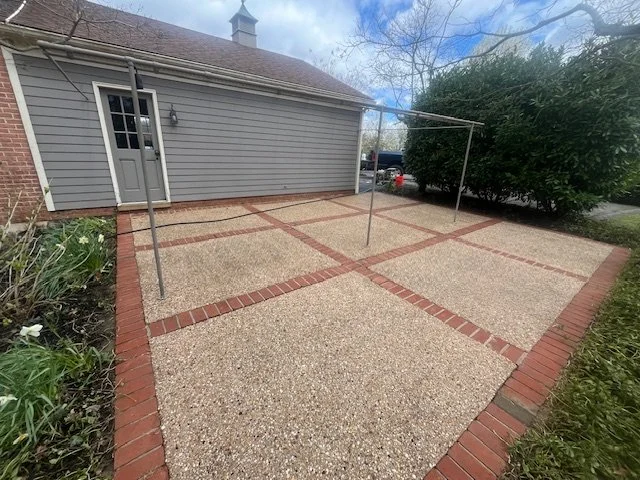 Backyard patio with gravel surface, brick edging, and a metal clothesline frame, next to a gray house with white trim and a door, and greenery on the sides.