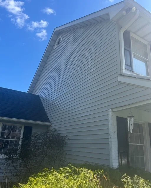 Close-up of the side of a white house with vinyl siding, a window, and part of a gabled roof, under a blue sky with a few clouds.