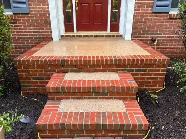 Brick front porch with two steps leading to a red front door, surrounded by shrubs and garden lights.