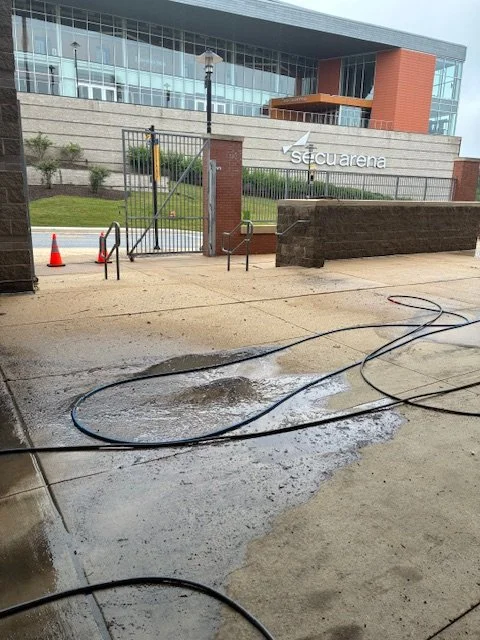 Wet sidewalk with hoses and water puddles outside the SecuArena building, which has glass windows and a brick and concrete exterior.