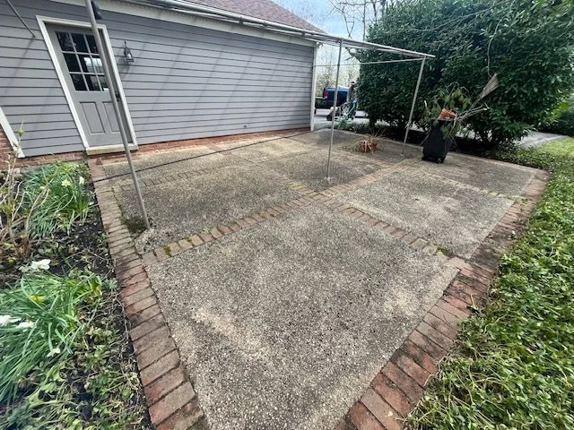 Empty backyard patio with concrete slabs, a brick border, and a clothesline frame, next to a house with gray siding and a door.
