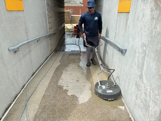 A man wearing a blue hat and long-sleeve shirt is operating a power floor buffer to clean a concrete pathway between two walls.