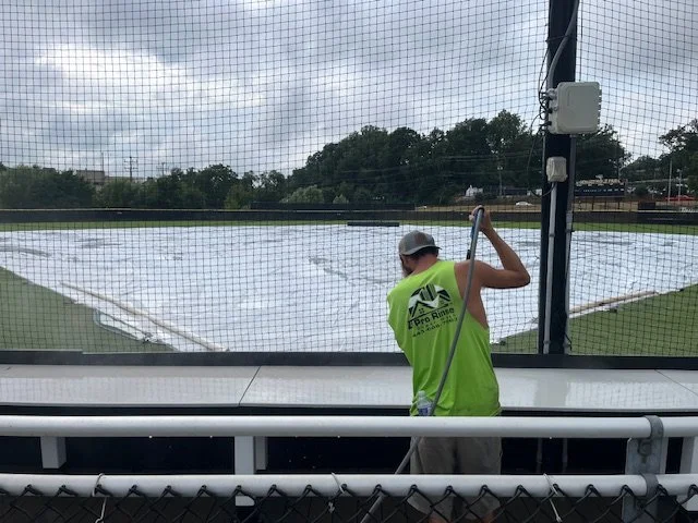 A worker with a neon vest watering a flooded outdoor sports field through a netted fence.
