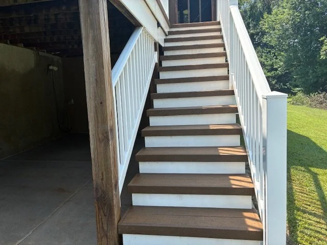 Exterior wooden staircase with white railings leading up to a deck, next to a grassy yard and trees in the background.