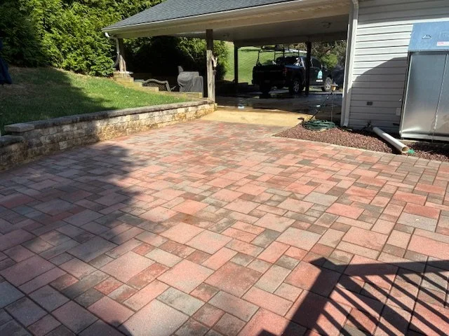 Paved backyard patio with red and gray bricks, adjacent to a house with a carport, and a shadow cast on the patio.