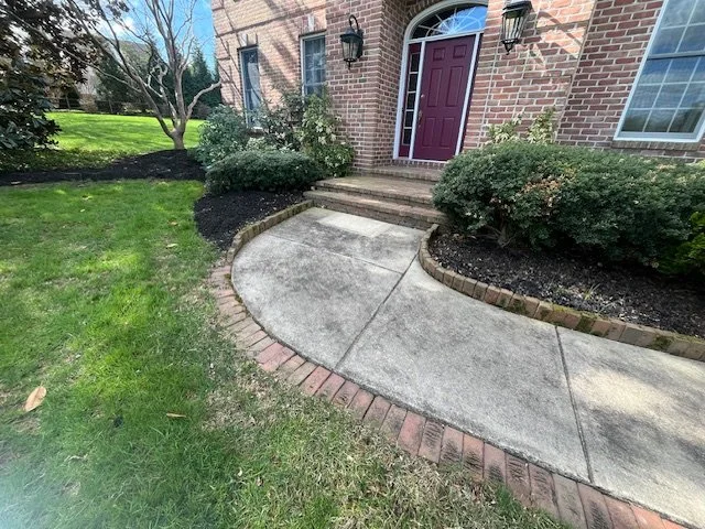 Concrete sidewalk leading to front door with brick steps, surrounded by bushes and mulch, in front of a brick house with a purple door.