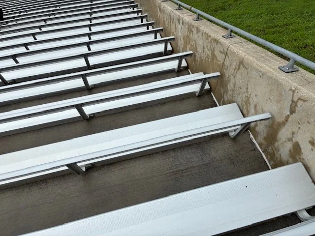 Empty metal bleacher seats on a concrete foundation next to a grassy area with a metal railing.